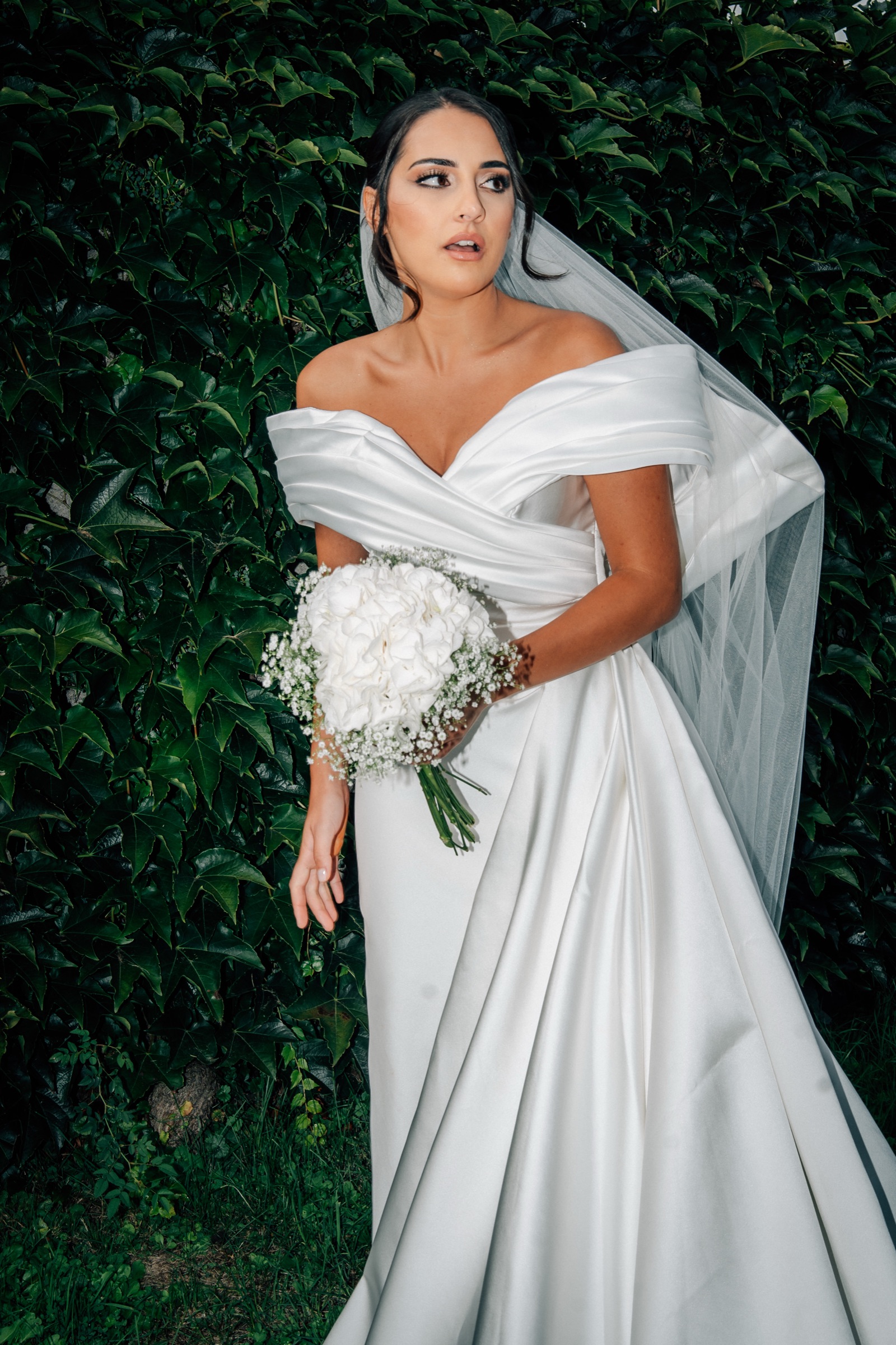 Braut im Off-Shoulder-Kleid mit Brautstrauß vor dunkler Hecke